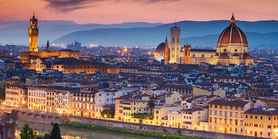 Panoramic view of Florence, Italy, highlighting the city’s iconic skyline with the Florence Cathedral (Duomo) and its red-tiled dome standing prominently. The Arno River flows through the city, in the foreground with the Tuscan hills in the background under a soft, golden sunset." 