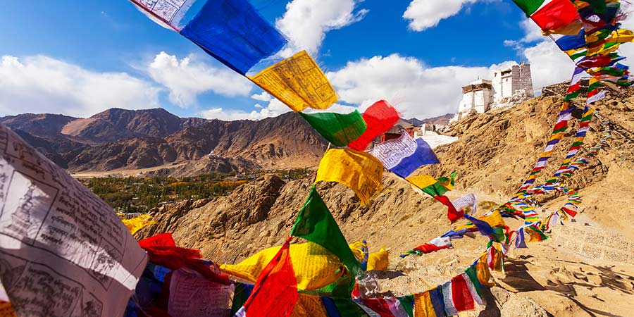 Image of Namgyal Monastery nestled on a hilltop with the rugged Himalayan mountains in the background. The monastery features traditional Tibetan architecture, with colourful prayer flags fluttering in the wind in the foreground. 